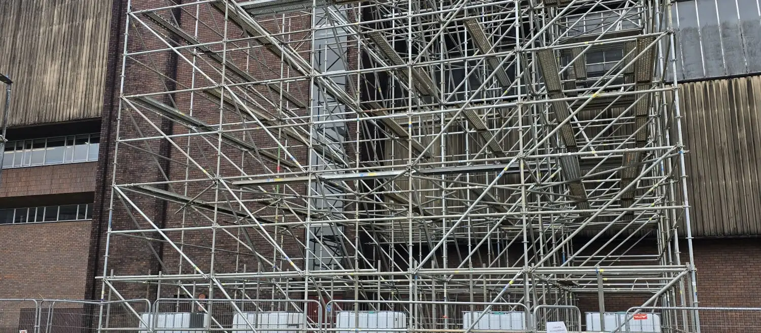Large scaffold structure against a tall industrial building, with a row of tanks at its base