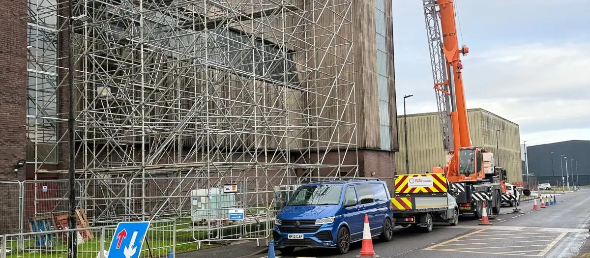 Full-height scaffolding across an industrial building’s façade, with a mobile crane and van on the road outside