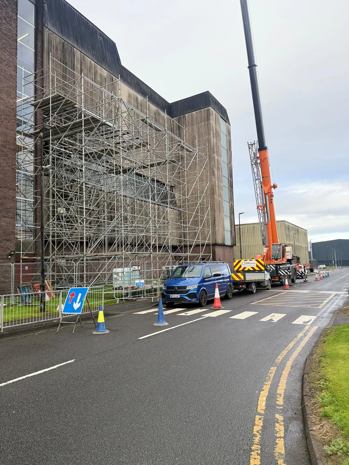 Full-height scaffolding across an industrial building’s façade, with a mobile crane and van on the road outside