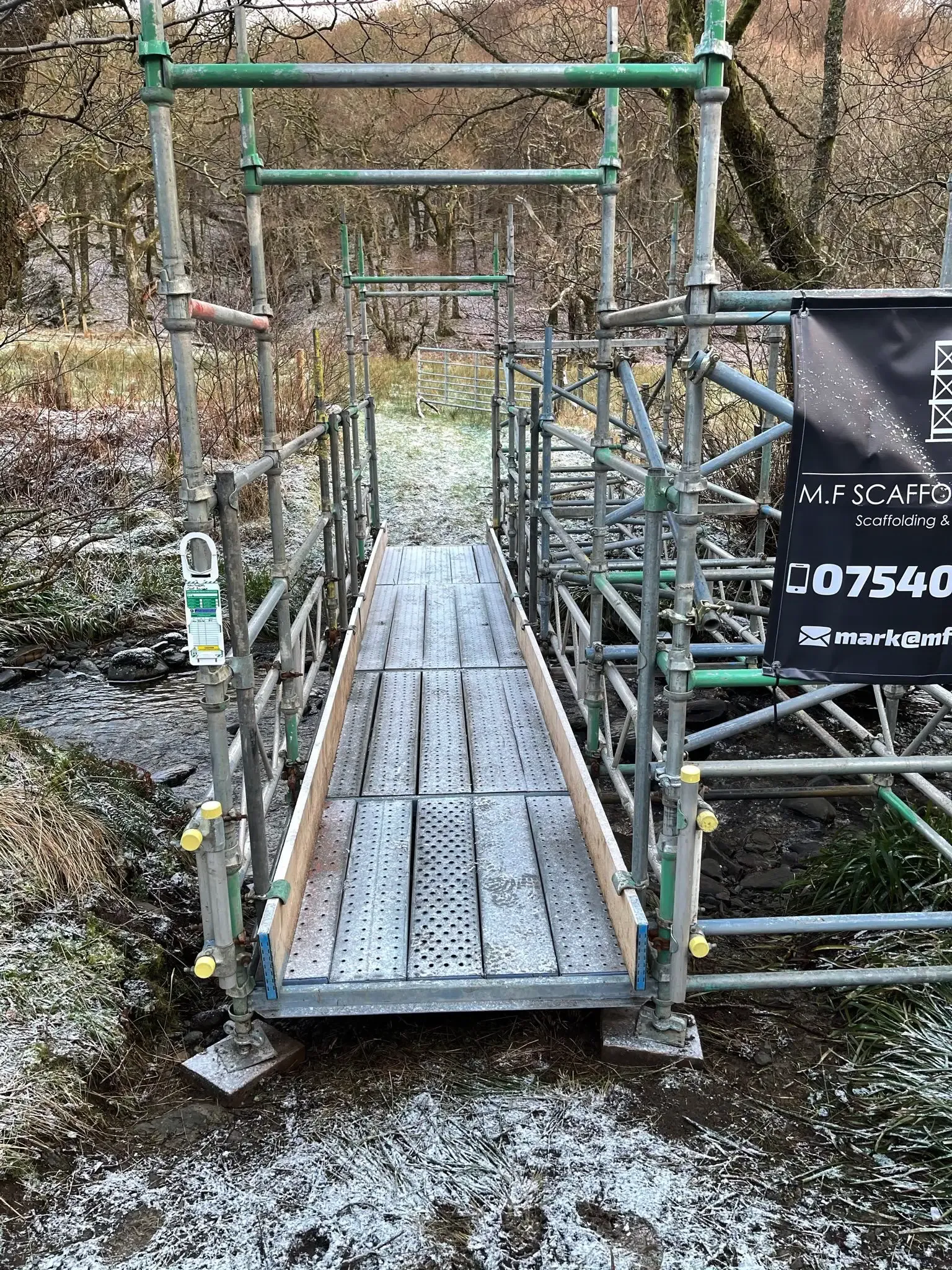 Scaffold footbridge with a steel deck and handrails over a burn