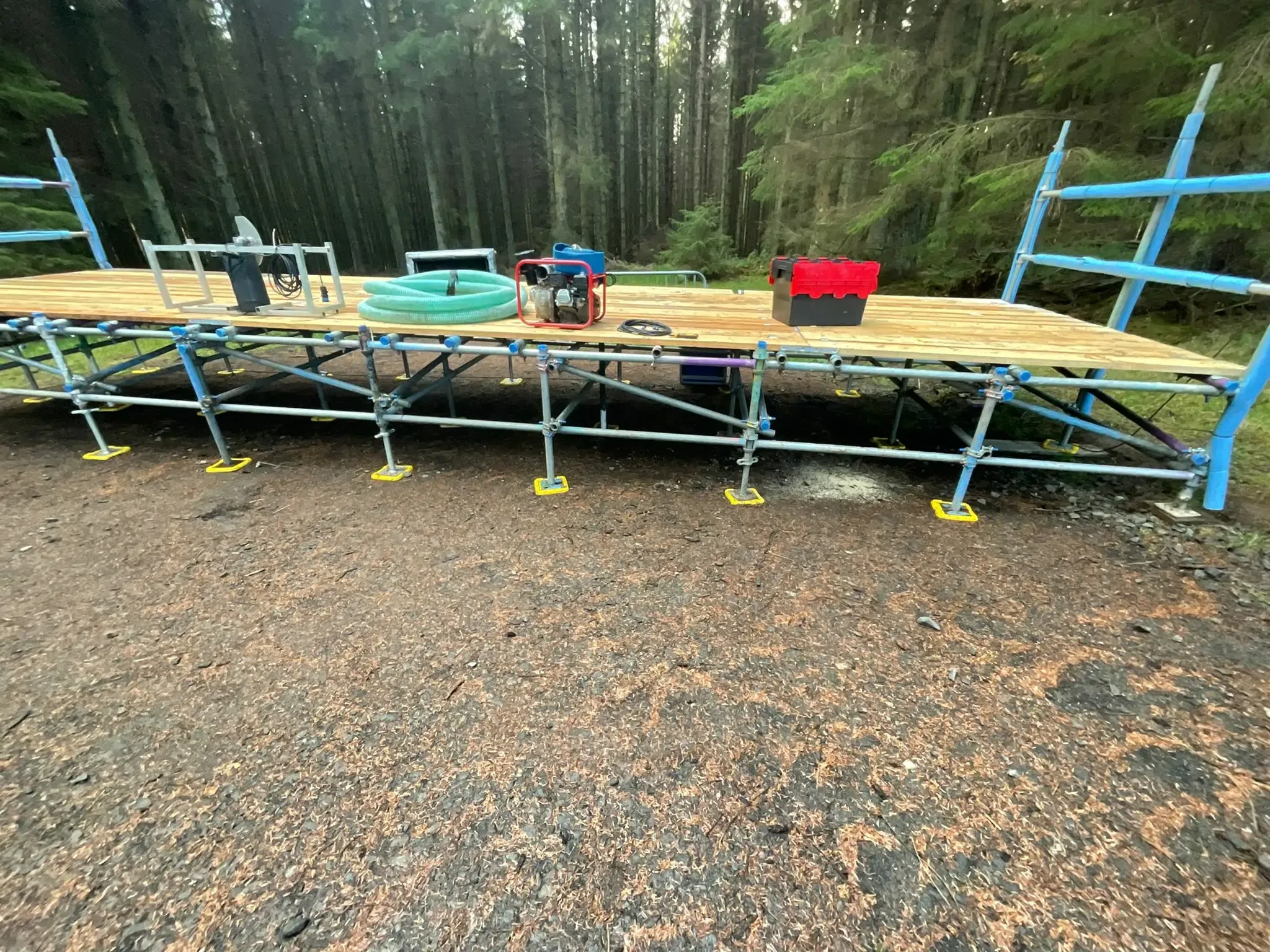 Boarded scaffold deck on adjustable bases over gravel, with woodland behind