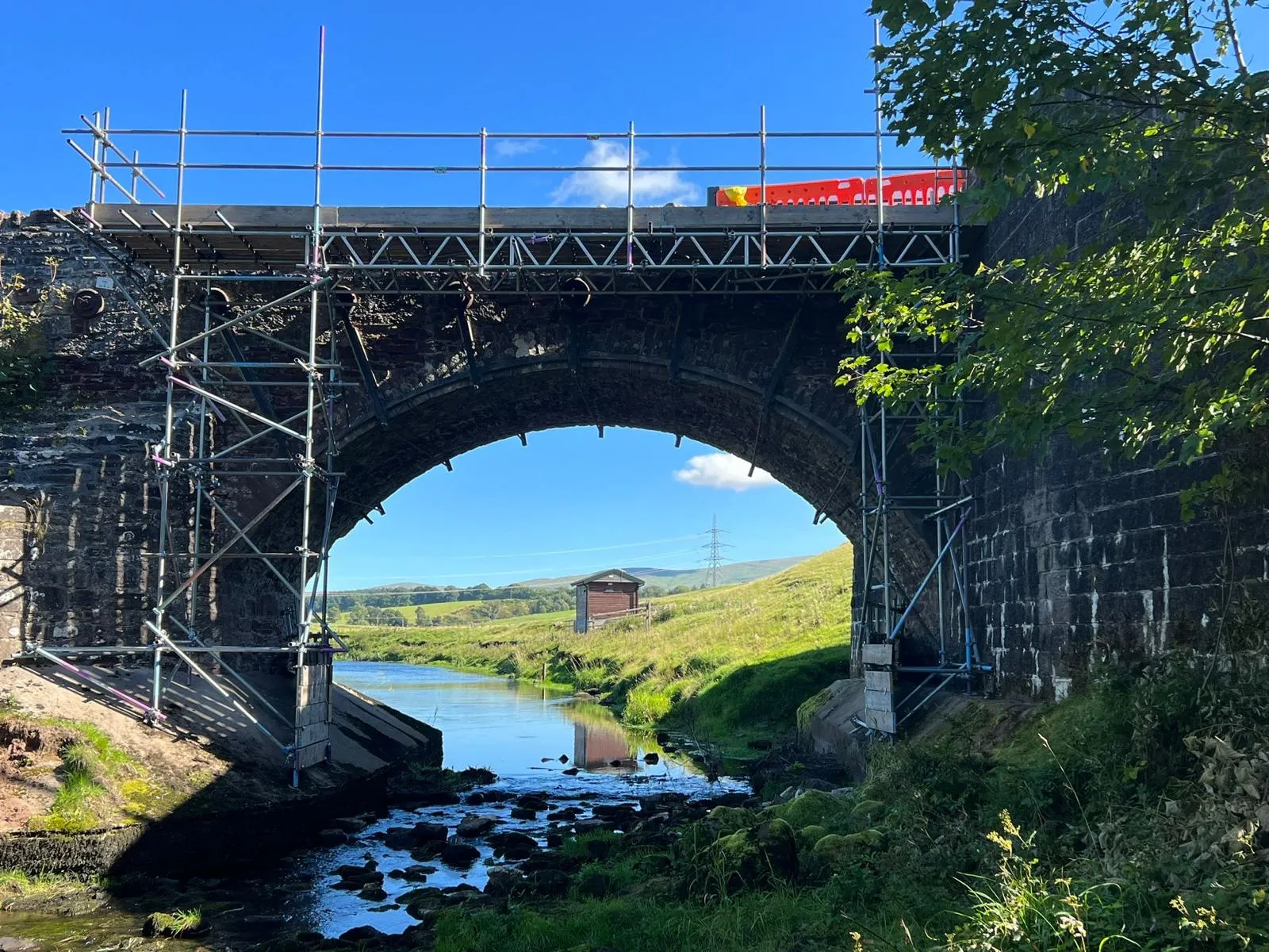 Scaffold with a beamed working platform across a stone arch bridge over a river